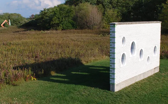 White wall with round windows stands in a grassy field, casting a long shadow.