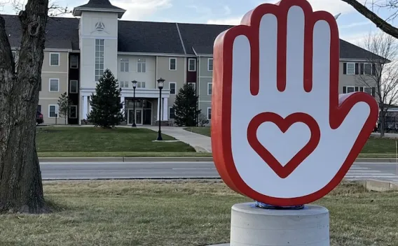 Red and white hand sculpture with a heart, in front of a large, light-colored building.