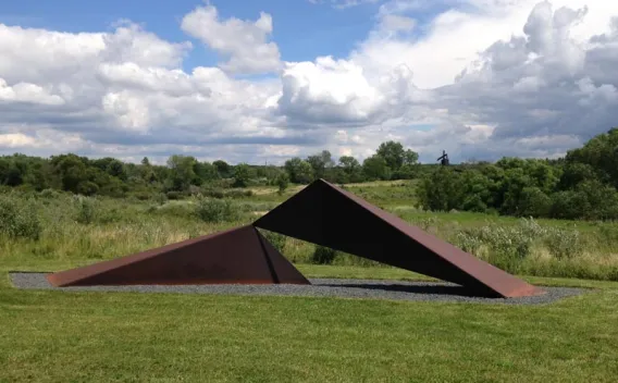 Large, rust-colored, angular metal sculpture in a green field with cloudy sky.