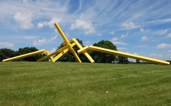Large yellow abstract sculpture on a green lawn under a blue sky with white clouds.