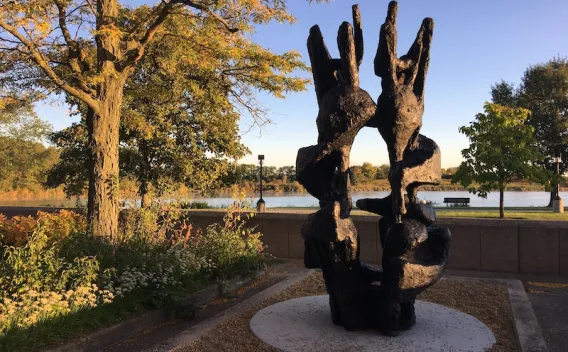 Photograph: Abstract dark sculpture by a river with autumn trees under a blue sky.
