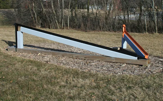 Gray and black angled ramp with colorful supports, on wood chips and grass.