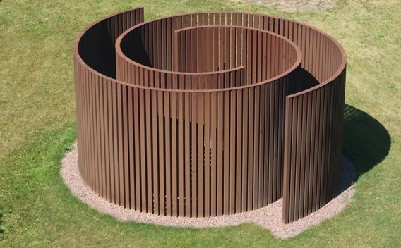Reddish-brown slatted spiral sculpture on a grassy lawn.