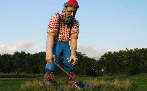 Large lumberjack statue with red hat, plaid shirt, holding an axe in a field.