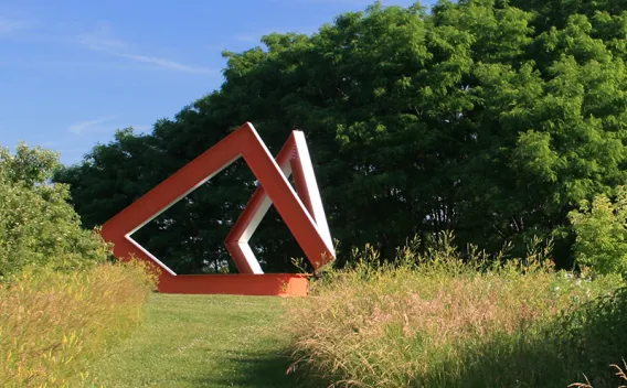 Red angular sculpture in a sunny green landscape.