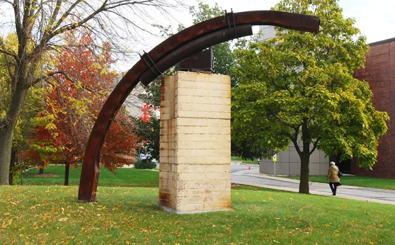 Outdoor sculpture: curved rusty metal beams over a light pedestal, autumn trees.