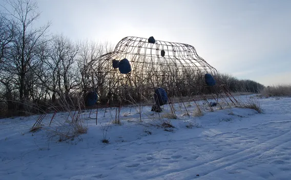 Woven branch dome on snowy hill with dark forms, sun glowing behind it.