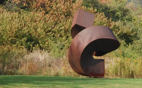 Large, rust-colored abstract metal sculpture in a green field with autumn trees.