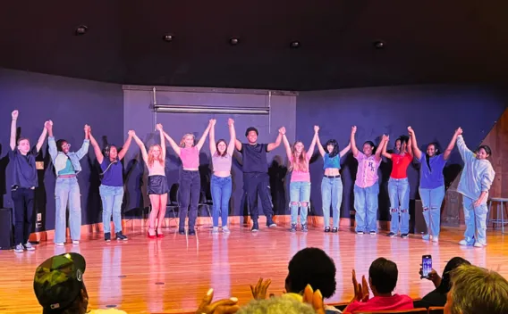 Diverse group on stage holding hands with raised arms for a curtain call, audience applauds.