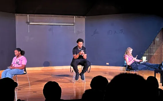 Three young actors on stage: two women in pink, one man in black, all looking down.