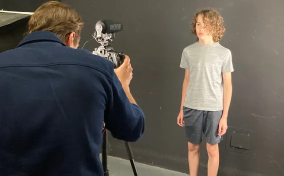 Photographer shoots a young boy standing against a dark studio background.