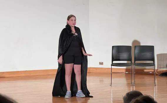 Teen performer in a long black cape, shorts, and sneakers stands on stage, arm extended.
