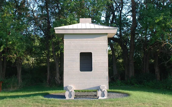 Modern light gray structure with dark window, on a gravel base, surrounded by trees.