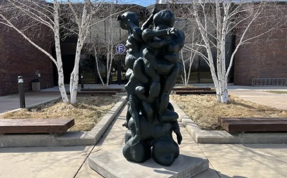 Dark abstract sculpture of stacked human forms outdoors, with a brick building and leafless trees.