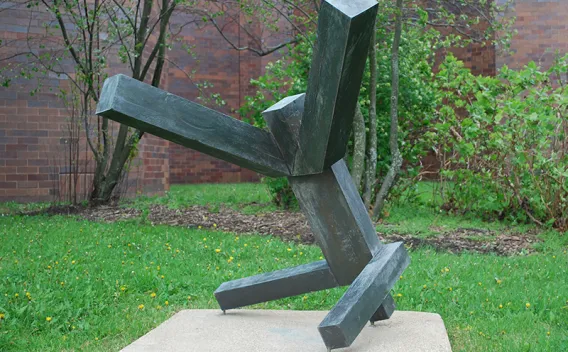 Geometric dark metal sculpture on a concrete base in a grassy outdoor setting.