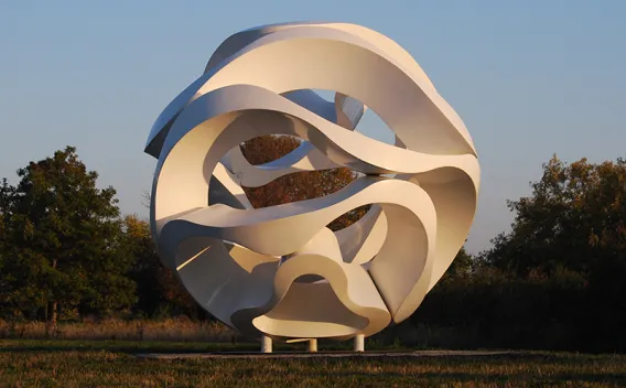 White, abstract, spherical sculpture with intertwining forms on a grassy field.