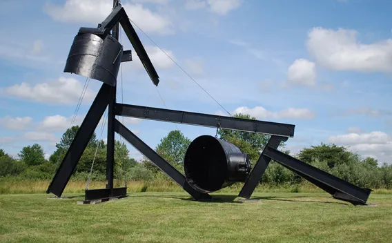 Large black abstract sculpture of industrial beams and cylinders in a green field.