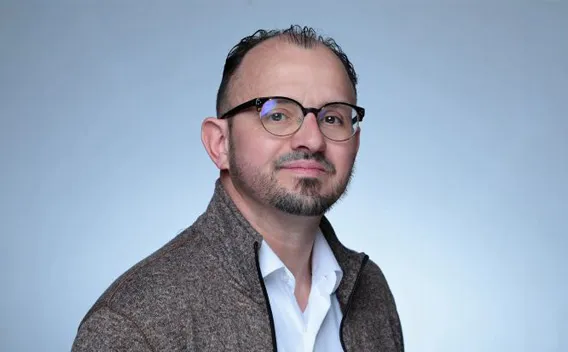 Man in glasses, with mustache and beard, wearing white shirt and brown jacket