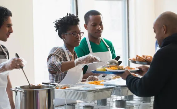 Volunteers serve hot food from large dishes to a person with a tray.