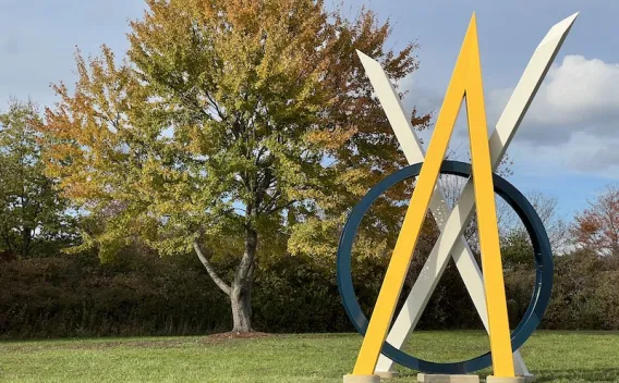 Geometric sculpture with blue, yellow, white shapes in a park, golden autumn tree.