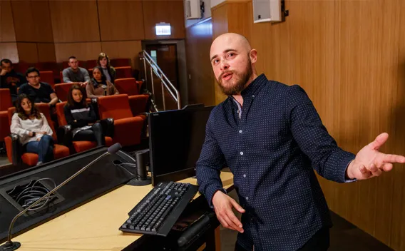 Bearded man in blue shirt gesturing at a podium in a lecture hall with students.