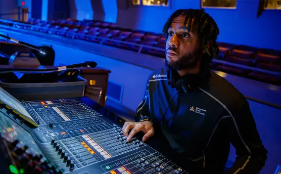 Man at soundboard in blue-lit auditorium, adjusting controls.