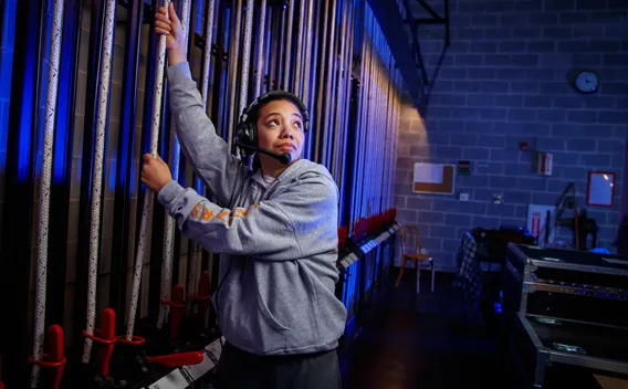Woman in headset pulling stage rigging backstage, blue lights.