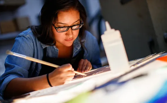 Young woman in glasses painting intently on a brightly lit table.