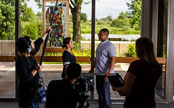 Photograph: Film crew records a man and woman, with boom mic and camera, near large windows.