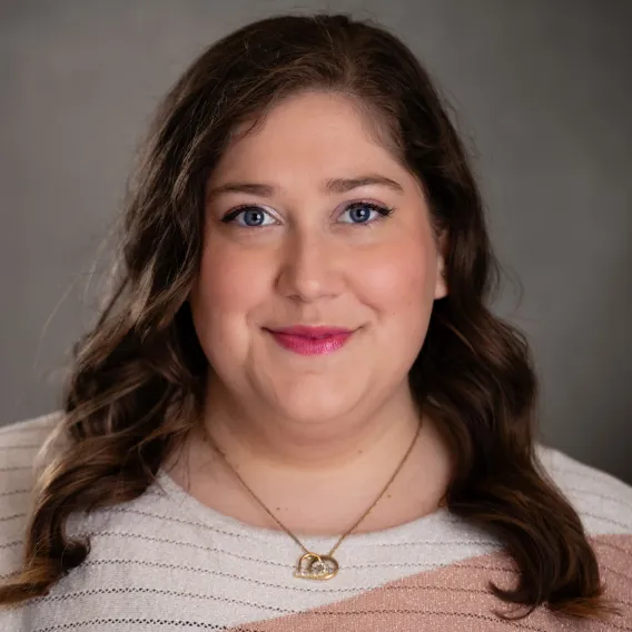 Person with long wavy hair wearing a light striped top and a gold necklace with interlocking rings, posed against a dark gray backdrop