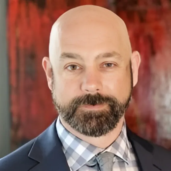 Person wearing a suit jacket, checkered shirt, and tie, posed in front of a red and dark-toned abstract background