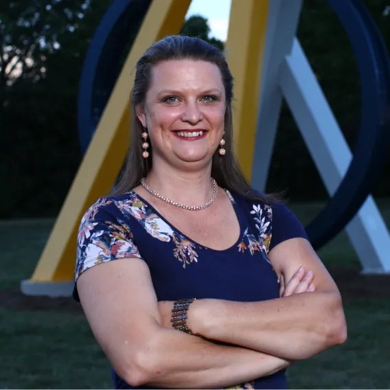 Person standing outdoors with arms crossed, wearing a floral navy top, with a large geometric metal sculpture in the background