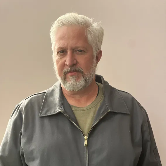 Person wearing a gray zip‑up jacket over a light shirt, standing in front of a plain beige background