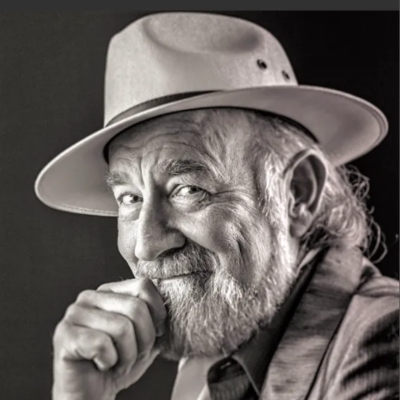  Person wearing a light-colored hat and suit jacket, posed in a dramatic black‑and‑white portrait with one hand resting near the chin