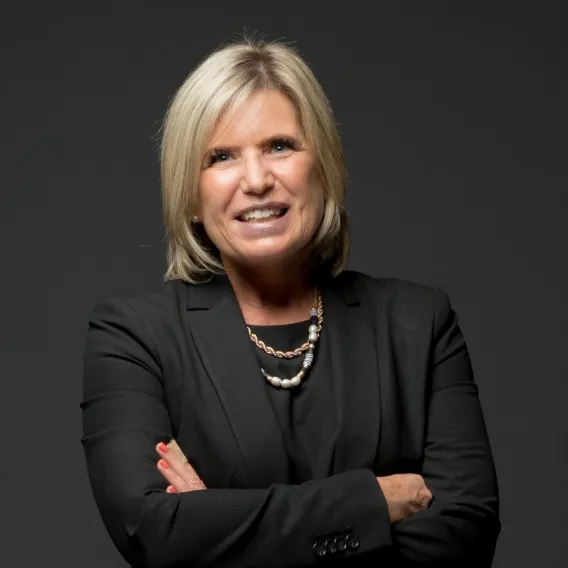 Person wearing a black blazer and layered necklace, standing with arms crossed against a dark background