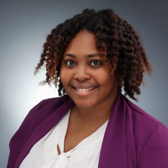 Person wearing a white blouse and a purple blazer, with short textured hair, posed against a smooth gray backdrop