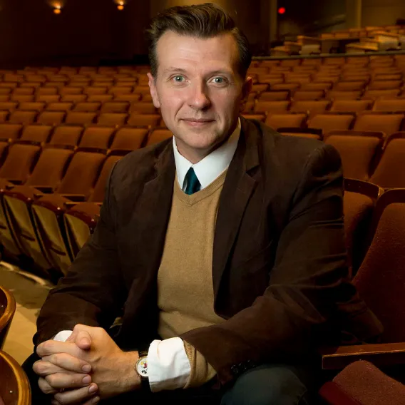 Person seated in an auditorium, wearing a brown jacket, tan sweater, and tie, with rows of empty theater seats in the background