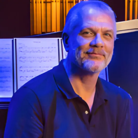 Person wearing a dark collared shirt seated beside an open music score, with stage lighting and warm illuminated panels in the background