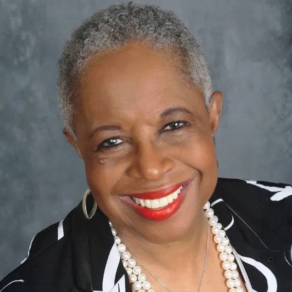  Person wearing a black and white patterned jacket and layered pearl necklaces, posed against a textured gray backdrop.