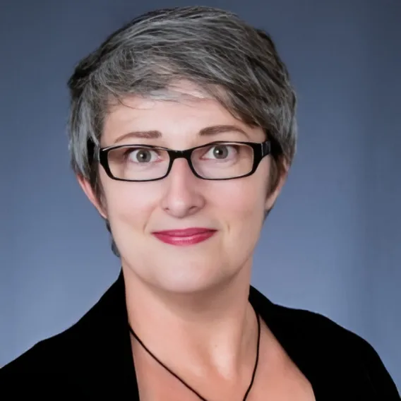 Person with short, gray hair wearing a black top and necklace, posed against a smooth blue‑gray backdrop