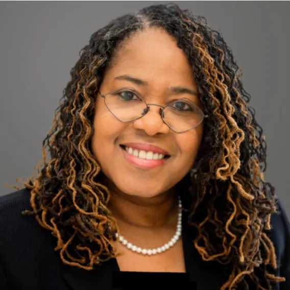 Person with long, curly hair wearing a black blazer and a pearl necklace, posed against a plain gray background.