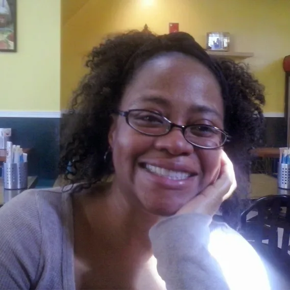 Person seated in a brightly lit café or restaurant, wearing a light gray top, with curly shoulder‑length hair and resting their head on one hand