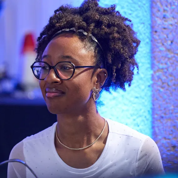 Person wearing a white shirt with subtle embellishments, sitting in front of a blue‑lit textured wall