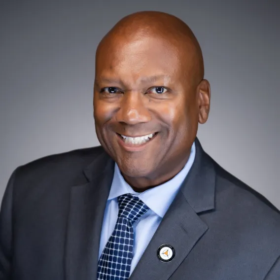  Person wearing a gray suit jacket, light blue shirt, and patterned blue tie, posed against a smooth gray background