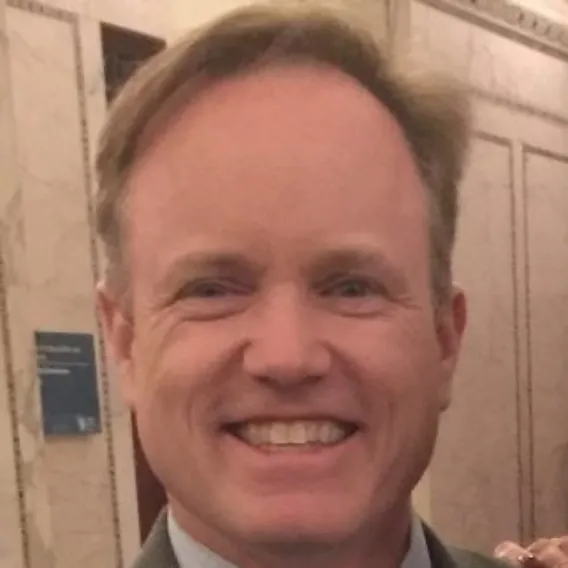 Person wearing a suit jacket and collared shirt standing in an ornate interior with marble walls and decorative architectural details