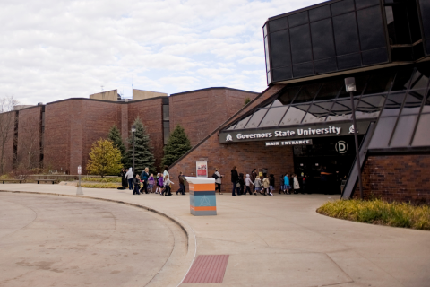 People walking toward the Governors State University main entrance, shown from the outside with brick buildings, landscaping, and a glass‑fronted entryway.